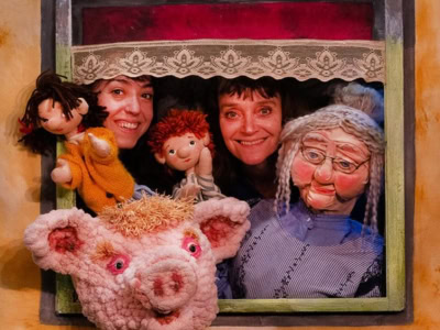 Two puppeteers smiling through a puppet theater window, holding colorful hand puppets including a girl in yellow, a boy with red hair, a gray-haired grandmother, and a fluffy pink pig. The scene is framed with bright colors and a lace curtain.