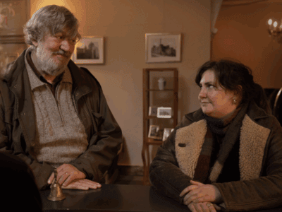 Still from Treasure: An older man (Stephen Fry) with a kind expression and a gray beard stands beside a woman (Lena Dunham) in a shearling coat at a hotel reception desk. They face a receptionist whose back is to the camera. The setting is warm and softly lit, suggesting a quiet, reflective moment.